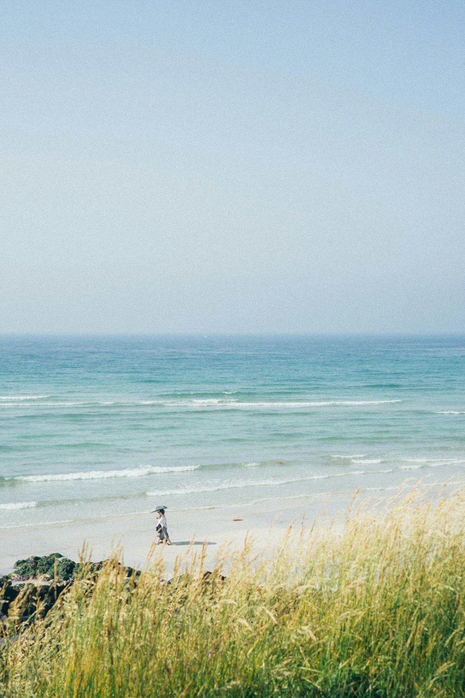 Person walks on a vast sandy beach beside gentle turquoise waves.