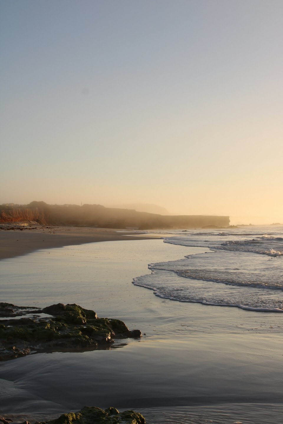 Gentle waves break against mossy rocks along a sandy beach.